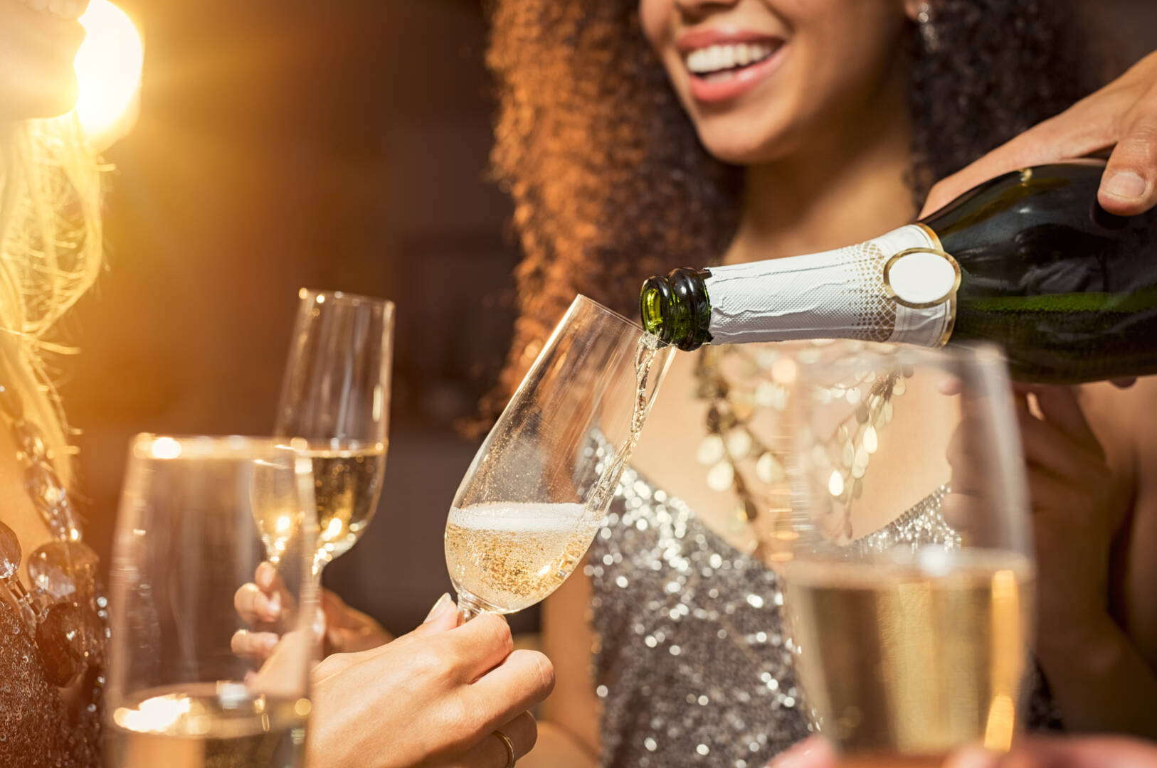 Etrafındaki arkadaşlarıyla birlikte şişeden bardaklara şampanya döken el. Parti sırasında flütlere beyaz şarap döken elin yakın çekimi. Yılbaşı kutlamasından detay görüntü.